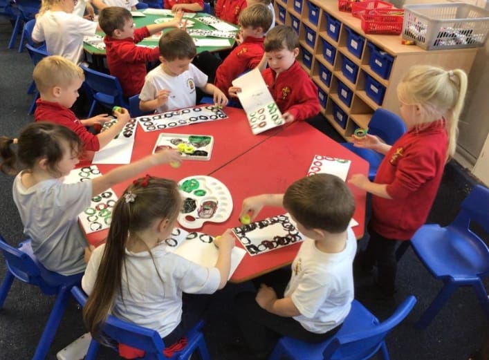 Primary pupils create colourful artwork together at a table using paint, palettes and printed shapes