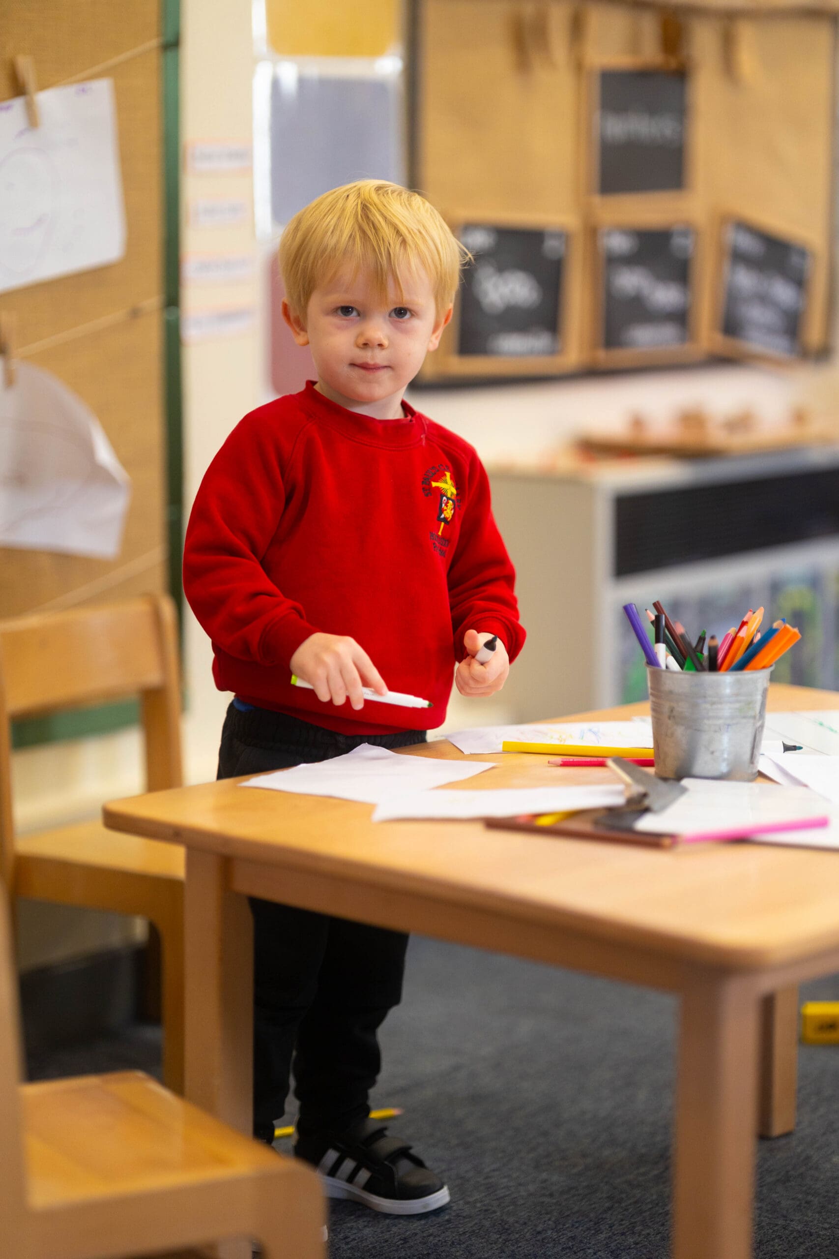 Young pupil in a red school jumper holding a whiteboard pen at a classroom table with drawing materials