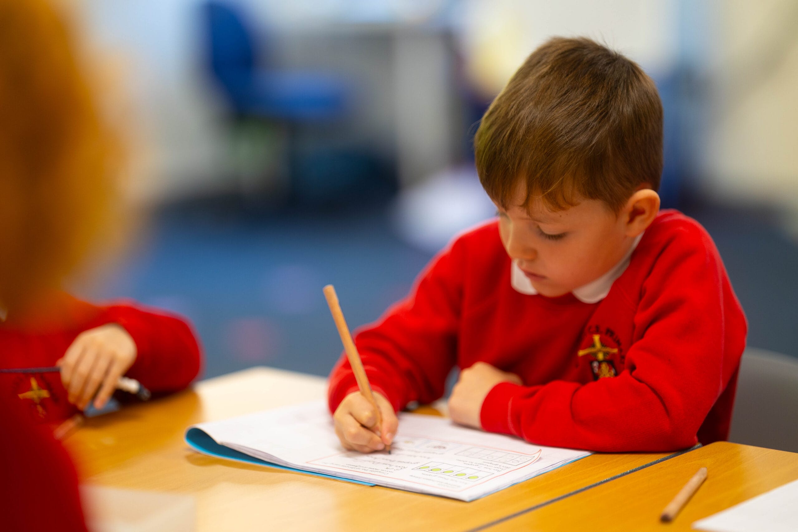 Pupil in a red school jumper writing in a workbook at a classroom table